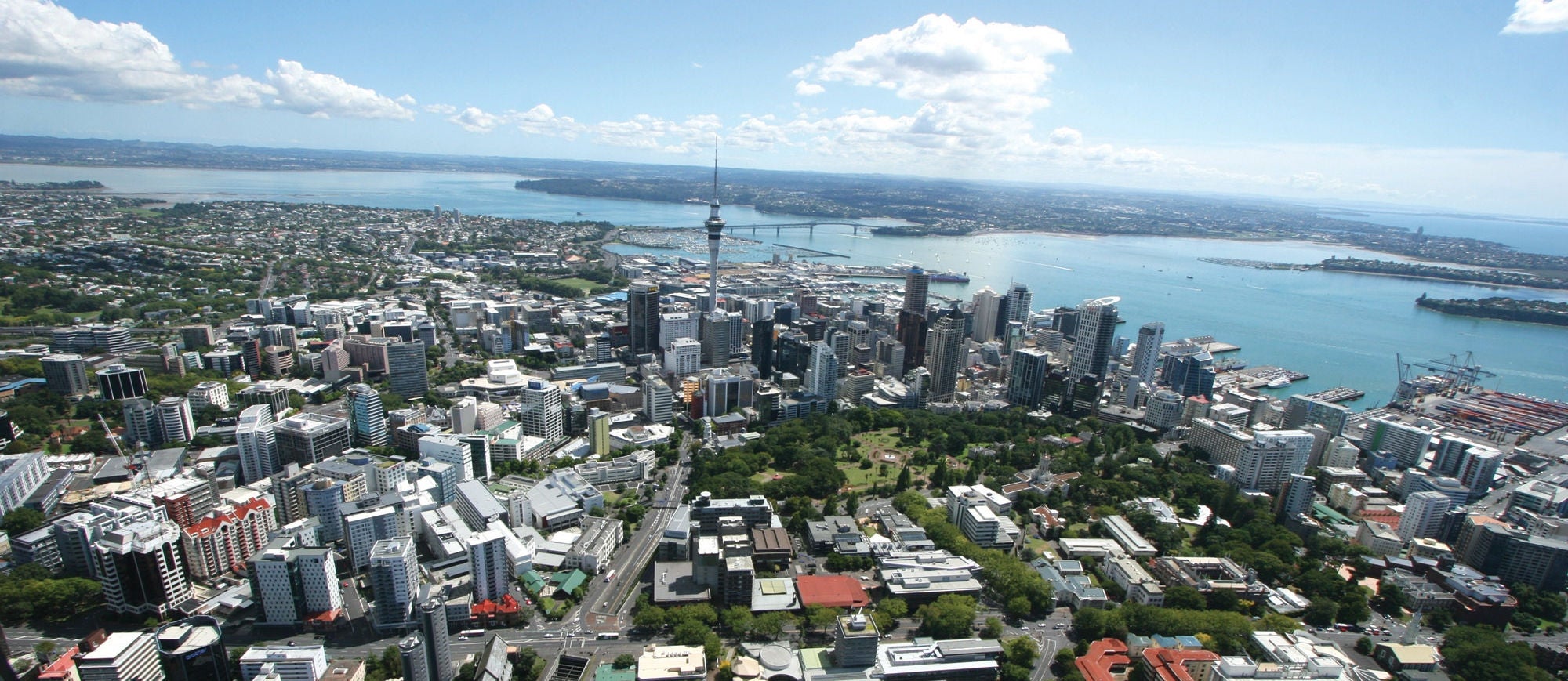Looking over Auckland. An elevated view looking north west over the Auckland CBD. It is a blue sky day with a few puffy clouds, and the skyscrapers of Auckland Tāmaki Makaurau shine in the morning sun, with the harbour bridge and north shore in the back ground.